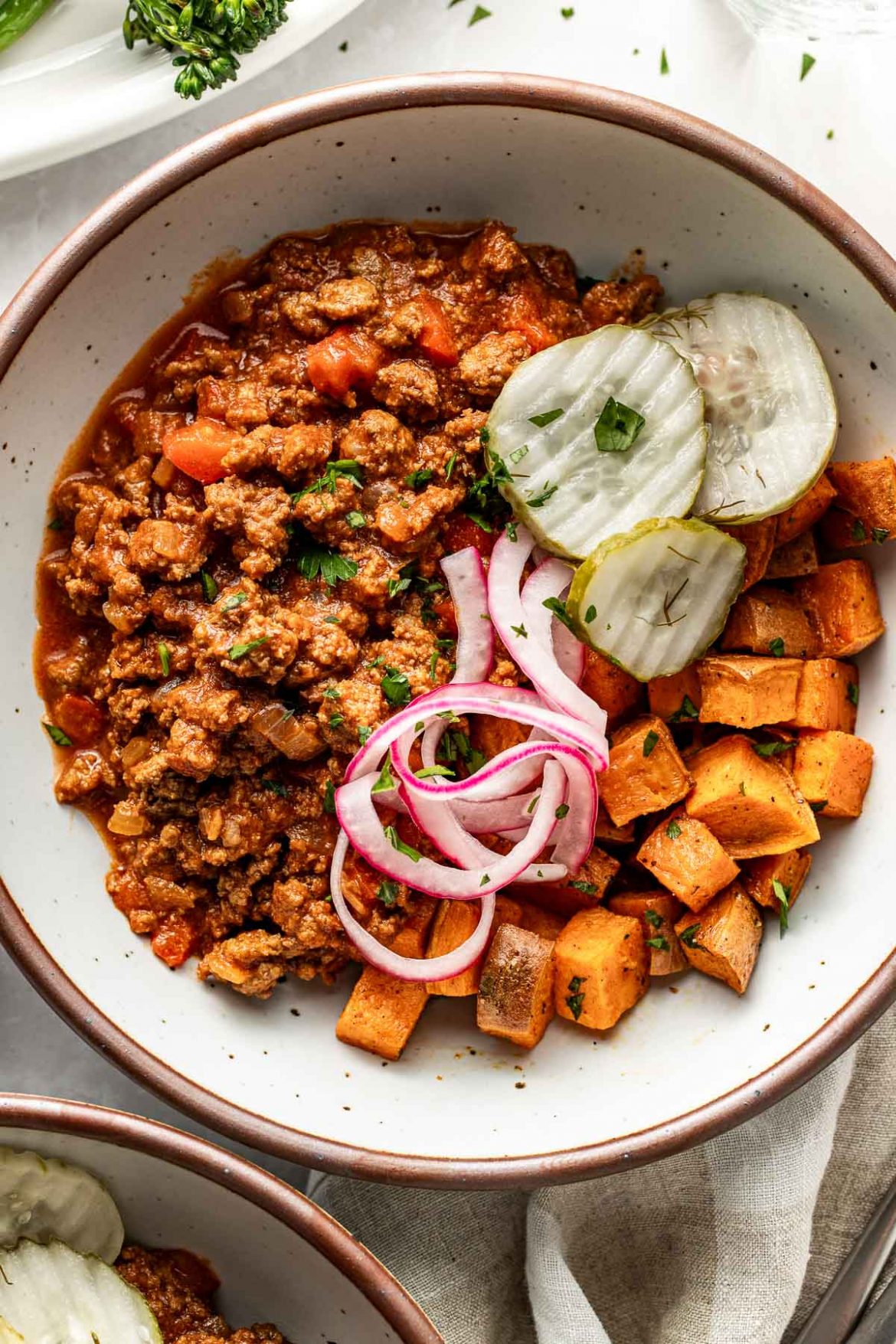 Overhead view of a bowl of sloppy joe meat, roasted sweet potatoes, pickled red onions, and crisp pickle chips. 